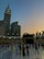 A view of the Kaaba at the Grand Mosque, surrounded by worshippers and pilgrims. The Abraj Al Bait Clock Tower is visible in the background, towering over the mosque complex. The sky is transitioning from day to night, with a blend of sunset colors.