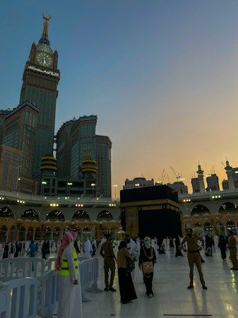 A view of the Kaaba at the Grand Mosque, surrounded by worshippers and pilgrims. The Abraj Al Bait Clock Tower is visible in the background, towering over the mosque complex. The sky is transitioning from day to night, with a blend of sunset colors.