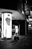 A black and white street scene at night featuring a brightly lit advertisement with a model wearing casual clothing. Below the advertisement, a person dressed in light-colored clothing is sitting on the curb and looking at their phone. In the background, there are several people walking and numerous illuminated store signs.