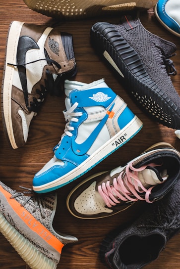 A collection of stylish sneakers arranged on a wooden floor. The footwear features various designs, including high-tops and low-tops, with a mix of materials such as leather and fabric. The color palette includes bright blues, neutral earth tones, and bold blacks, with some sneakers showcasing unique branding details.