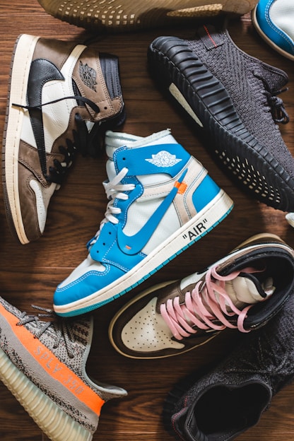 A collection of stylish sneakers arranged on a wooden floor. The footwear features various designs, including high-tops and low-tops, with a mix of materials such as leather and fabric. The color palette includes bright blues, neutral earth tones, and bold blacks, with some sneakers showcasing unique branding details.