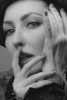 A monochrome portrait of a person with expressive eyes and delicate features. The individual is adorned in stylish clothing, partially covered by a hat. Their hand is elegantly posed near their face, partially covering their lips.