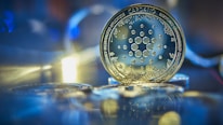 A close-up view of a Cardano cryptocurrency coin, surrounded by a soft-focus background with bokeh effects and reflections. The coin displays intricate circuit-like patterns, suggesting advanced technology.