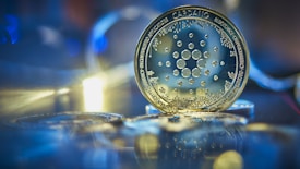 A close-up view of a Cardano cryptocurrency coin, surrounded by a soft-focus background with bokeh effects and reflections. The coin displays intricate circuit-like patterns, suggesting advanced technology.