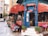 A charming street-side cafe with red awnings and a blue entrance, surrounded by potted plants. There are tables with colorful tablecloths set up along the sidewalk. The cafe offers a menu displayed on a chalkboard with items such as breakfast, lunch, and dessert. People are seen inside and outside, giving it a lively and inviting atmosphere.