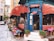 A charming street-side cafe with red awnings and a blue entrance, surrounded by potted plants. There are tables with colorful tablecloths set up along the sidewalk. The cafe offers a menu displayed on a chalkboard with items such as breakfast, lunch, and dessert. People are seen inside and outside, giving it a lively and inviting atmosphere.
