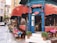 A charming street-side cafe with red awnings and a blue entrance, surrounded by potted plants. There are tables with colorful tablecloths set up along the sidewalk. The cafe offers a menu displayed on a chalkboard with items such as breakfast, lunch, and dessert. People are seen inside and outside, giving it a lively and inviting atmosphere.