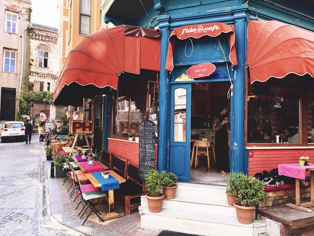 A charming street-side cafe with red awnings and a blue entrance, surrounded by potted plants. There are tables with colorful tablecloths set up along the sidewalk. The cafe offers a menu displayed on a chalkboard with items such as breakfast, lunch, and dessert. People are seen inside and outside, giving it a lively and inviting atmosphere.