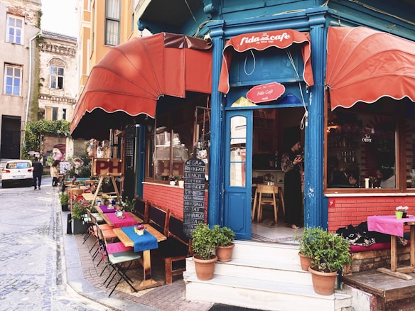 A charming street-side cafe with red awnings and a blue entrance, surrounded by potted plants. There are tables with colorful tablecloths set up along the sidewalk. The cafe offers a menu displayed on a chalkboard with items such as breakfast, lunch, and dessert. People are seen inside and outside, giving it a lively and inviting atmosphere.