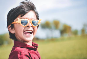 A cheerful toddler wearing colorful round glasses, smiling outdoors.