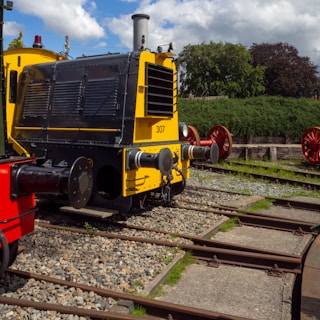 A bright yellow and black train engine is parked on tracks surrounded by gravel and greenery. The engine has prominent front buffers, and a red-painted component is visible along its side. The scene is set outdoors under a partly cloudy sky.