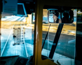 View from a bus driver's cabin with a rearview mirror reflecting a person wearing a hat. The interior consists of various dashboard elements and controls. Through the front windshield, tram tracks and a blurred background create a sense of motion.