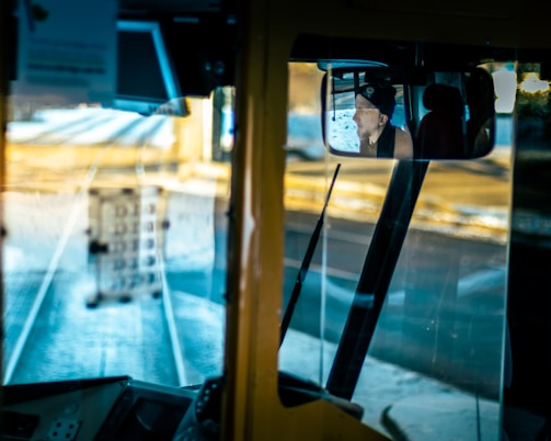 View from a bus driver's cabin with a rearview mirror reflecting a person wearing a hat. The interior consists of various dashboard elements and controls. Through the front windshield, tram tracks and a blurred background create a sense of motion.
