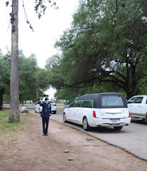 A uniformed person is saluting on a path near a road. A white hearse and other vehicles are parked nearby, surrounded by trees and greenery. The atmosphere is solemn and respectful.