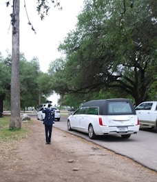 A uniformed person is saluting on a path near a road. A white hearse and other vehicles are parked nearby, surrounded by trees and greenery. The atmosphere is solemn and respectful.