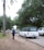 A uniformed person is saluting on a path near a road. A white hearse and other vehicles are parked nearby, surrounded by trees and greenery. The atmosphere is solemn and respectful.