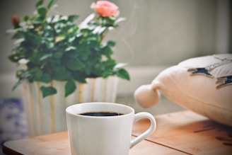 A steaming cup of black coffee sits on a wooden table next to a white pillow with decorative stitching. In the background, a green potted plant with pink flowers adds a touch of color.