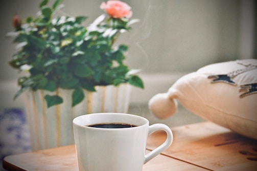 A steaming cup of black coffee sits on a wooden table next to a white pillow with decorative stitching. In the background, a green potted plant with pink flowers adds a touch of color.