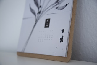 A close-up of a minimalist calendar with artistic black ink illustrations and date numbers, mounted on a thin wooden board. The calendar features abstract shapes and a monochromatic color scheme, emphasizing simplicity and elegance.