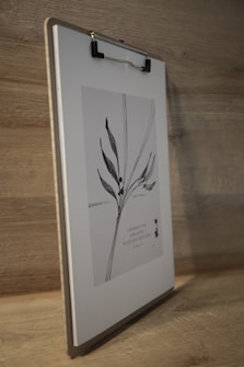 A wooden surface holds an upright clipboard with a botanical illustration. The illustration features delicate black leaves and stems against a light background. Below the illustration, there's text in a serif font.
