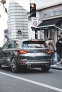 A black SUV is stationary at a traffic light in an urban setting. The rear of the vehicle has a license plate and is near a building with a store sign labeled 'Optic St. Honoré'. Pedestrians wearing coats and one with a face mask are visible on the sidewalk next to the store. A traffic sign indicates a speed limit of 30. The architecture in the background is typical of European cityscapes.