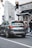 A black SUV is stationary at a traffic light in an urban setting. The rear of the vehicle has a license plate and is near a building with a store sign labeled 'Optic St. Honoré'. Pedestrians wearing coats and one with a face mask are visible on the sidewalk next to the store. A traffic sign indicates a speed limit of 30. The architecture in the background is typical of European cityscapes.