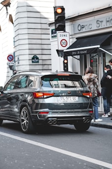 A black SUV is stationary at a traffic light in an urban setting. The rear of the vehicle has a license plate and is near a building with a store sign labeled 'Optic St. Honoré'. Pedestrians wearing coats and one with a face mask are visible on the sidewalk next to the store. A traffic sign indicates a speed limit of 30. The architecture in the background is typical of European cityscapes.