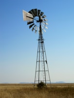 A windmill standing tall in an open field with blue skies, powering an industrial site.