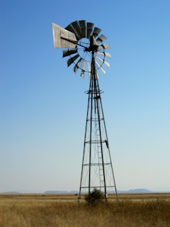 A windmill standing tall in an open field with blue skies, powering an industrial site.