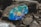 A large rock features a vibrant, colorful painting of a cartoon character. The rock is surrounded by other gray and brown rocks, creating a natural, rugged setting. Various doodles in bright colors like blue, orange, and purple decorate the surface.