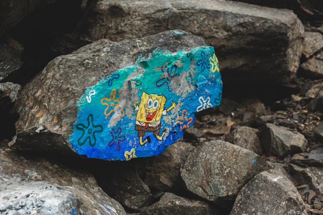 A large rock features a vibrant, colorful painting of a cartoon character. The rock is surrounded by other gray and brown rocks, creating a natural, rugged setting. Various doodles in bright colors like blue, orange, and purple decorate the surface.