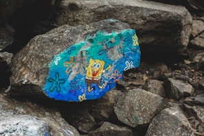 A large rock features a vibrant, colorful painting of a cartoon character. The rock is surrounded by other gray and brown rocks, creating a natural, rugged setting. Various doodles in bright colors like blue, orange, and purple decorate the surface.