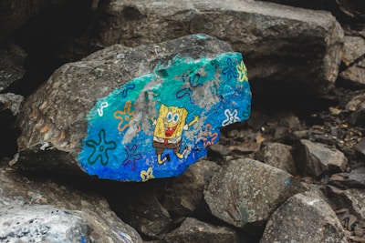 A large rock features a vibrant, colorful painting of a cartoon character. The rock is surrounded by other gray and brown rocks, creating a natural, rugged setting. Various doodles in bright colors like blue, orange, and purple decorate the surface.