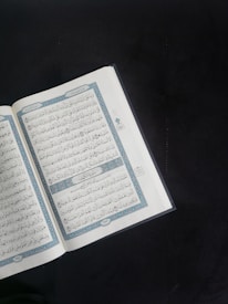 An open book with Arabic script is displayed prominently against a dark background. The pages are adorned with intricate blue and red decorations framing the text.