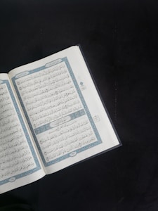 An open book with Arabic script is displayed prominently against a dark background. The pages are adorned with intricate blue and red decorations framing the text.