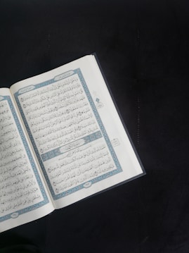 An open book with Arabic script is displayed prominently against a dark background. The pages are adorned with intricate blue and red decorations framing the text.