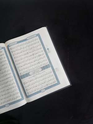 An open book with Arabic script is displayed prominently against a dark background. The pages are adorned with intricate blue and red decorations framing the text.
