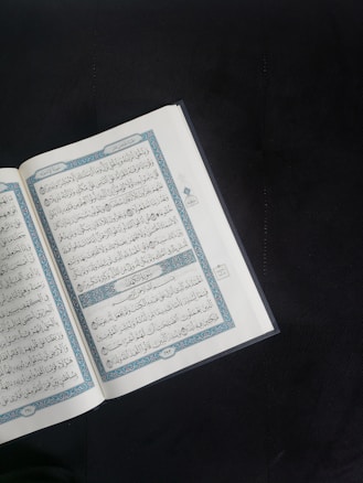 An open book with Arabic script is displayed prominently against a dark background. The pages are adorned with intricate blue and red decorations framing the text.