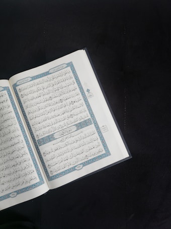 An open book with Arabic script is displayed prominently against a dark background. The pages are adorned with intricate blue and red decorations framing the text.