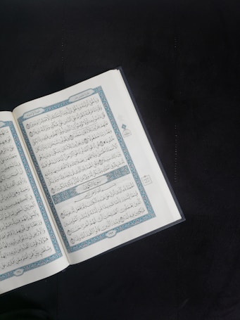 An open book with Arabic script is displayed prominently against a dark background. The pages are adorned with intricate blue and red decorations framing the text.