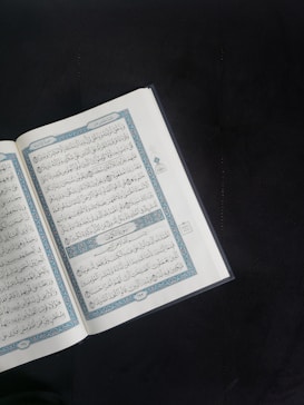 An open book with Arabic script is displayed prominently against a dark background. The pages are adorned with intricate blue and red decorations framing the text.