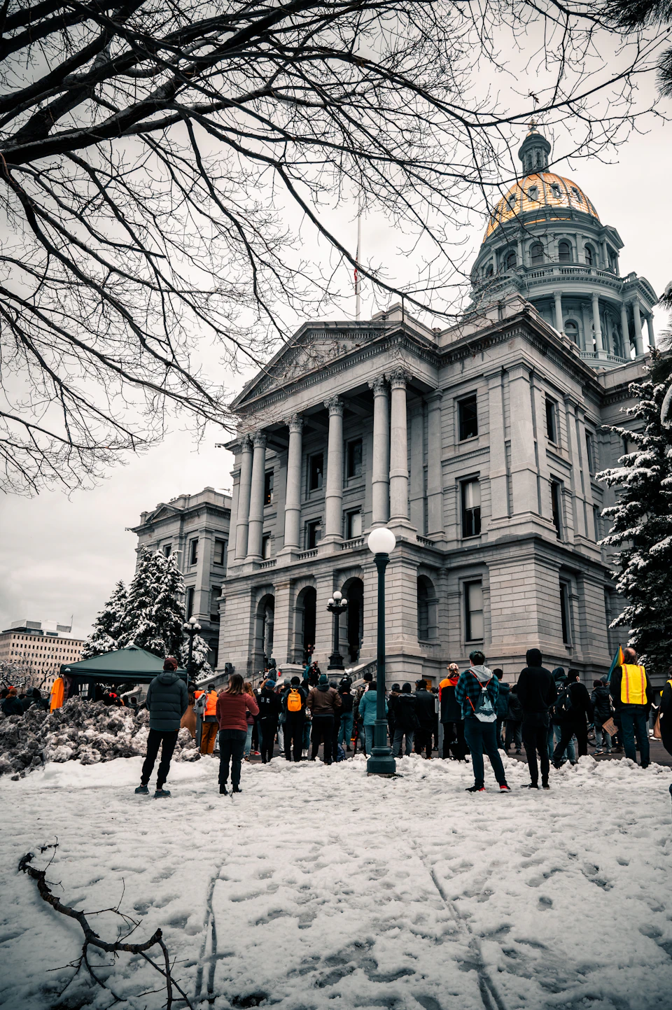 The image depicts a large, ornate building with a golden dome and columns, standing tall against an overcast sky. The building is surrounded by trees, adding to its majestic appearance. A group of people can be seen gathered outside the building, some carrying backpacks and one person holding a camera. The scene suggests that they might be tourists or visitors admiring the architectural beauty of the structure.