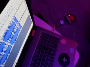 A laptop with waveforms displayed on the screen, suggesting audio editing or production. The desk is illuminated by purple lighting, and there are two stickers on the laptop. In the background, a small device with glowing red lights is visible on the carpeted floor.