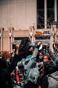 A group of people gathered in a protest, raising their fists in solidarity. One person holds a megaphone, indicating a leadership role or involvement in addressing the crowd. The scene takes place in an urban environment with a building and some leafless trees visible in the background.