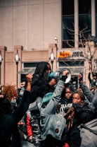 A group of people gathered in a protest, raising their fists in solidarity. One person holds a megaphone, indicating a leadership role or involvement in addressing the crowd. The scene takes place in an urban environment with a building and some leafless trees visible in the background.