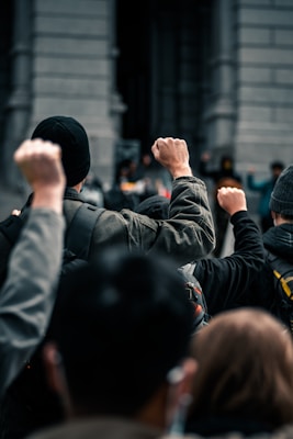 A group of people stand with raised fists, symbolizing unity and protest. They are outdoors, with the background showing a blurred structure that looks like an urban or governmental building.