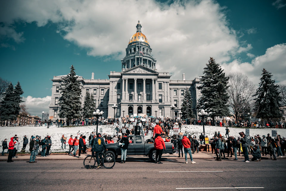 The image depicts a large crowd gathered outside the historic Fort Collins State Capitol building in Colorado, USA. The Capitol is a grand white structure with a gold dome and columns that stands tall against the sky. The people are standing on a street corner, some holding signs and banners, suggesting they may be participating in a protest or rally.

In front of the Capitol, there's a black truck parked, possibly belonging to one of the protesters. A few bicycles can also be seen among the crowd, indicating that some attendees chose eco-friendly transportation for their journey. The sky above is blue with scattered clouds, creating a peaceful and serene atmosphere around the historic building.