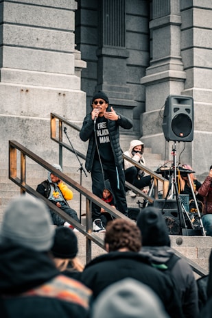 A person stands on a set of stone steps, holding a microphone and wearing a black jacket and hat. The individual is addressing a crowd, as other people sit or stand nearby, some with masks or headphones. A speaker and microphone stand are also visible, along with a mix of attendees in casual attire.