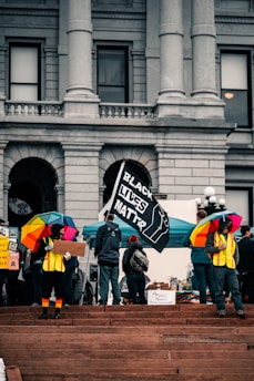A group of people is gathered on the steps outside a large building with archways and columns. Several individuals are holding umbrellas with rainbow patterns, and one person is holding a 'Black Lives Matter' flag. Another person is holding a cardboard sign. The scene appears to be a protest or demonstration.