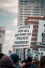 Protestors are gathered holding signs demanding justice for victims of police violence. The main sign in focus reads 'Justice for all victims of police terror!' with a cityscape in the background.